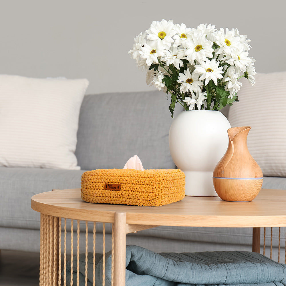 Coffee table with a vase of white flowers, wooden diffuser, and yellow tissue box in a living room setting.