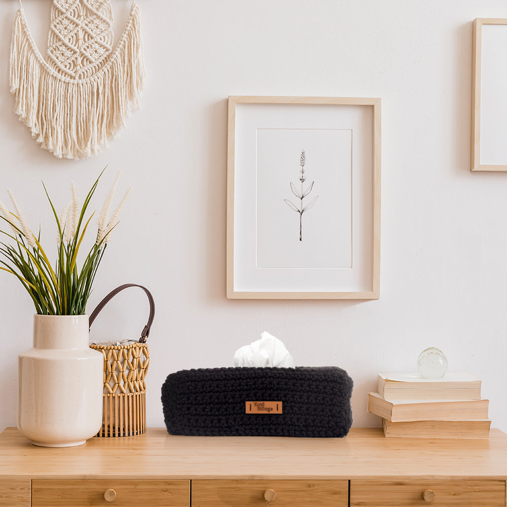 Decorative items on a wooden surface including a black tissue box, books, a plant, and a macrame wall hanging.