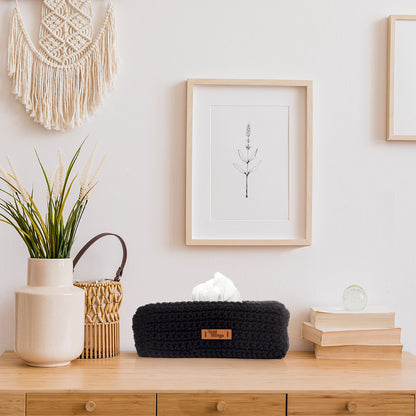 Decorative items on a wooden surface including a black tissue box, books, a plant, and a macrame wall hanging.
