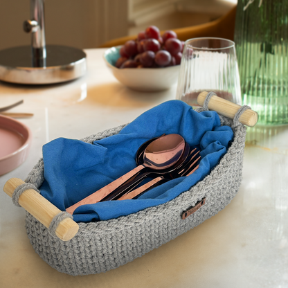 Gray woven basket with wooden handles containing cutlery on a kitchen counter.
