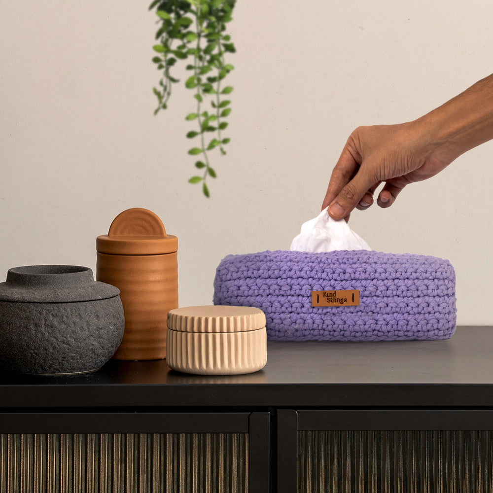 Hand reaching for a tissue from a purple woven tissue holder on a table with decorative jars.