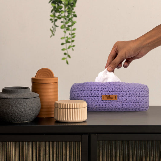 Hand reaching for a tissue from a purple woven tissue holder on a table with decorative jars.