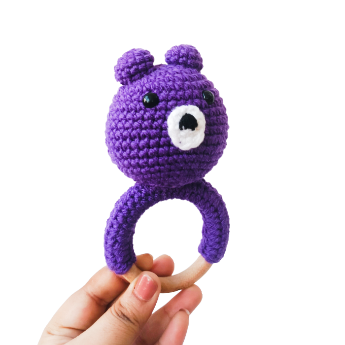 Purple crochet bear rattle held by a hand on a white background