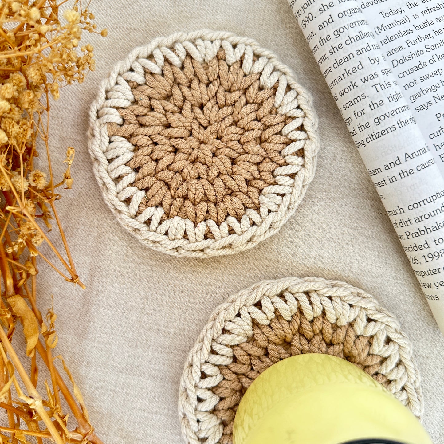 Two crocheted coasters on a textured surface with an open book and dried plants.