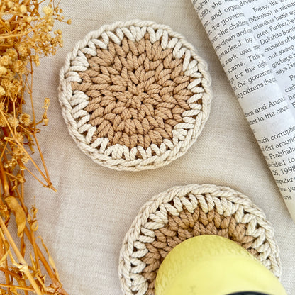 Two crocheted coasters on a textured surface with an open book and dried plants.