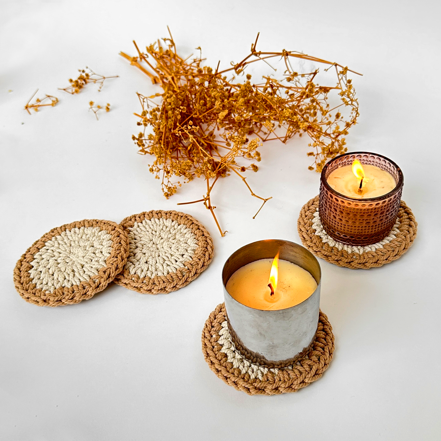 Two lit candles on woven coasters with dried plants on a white background